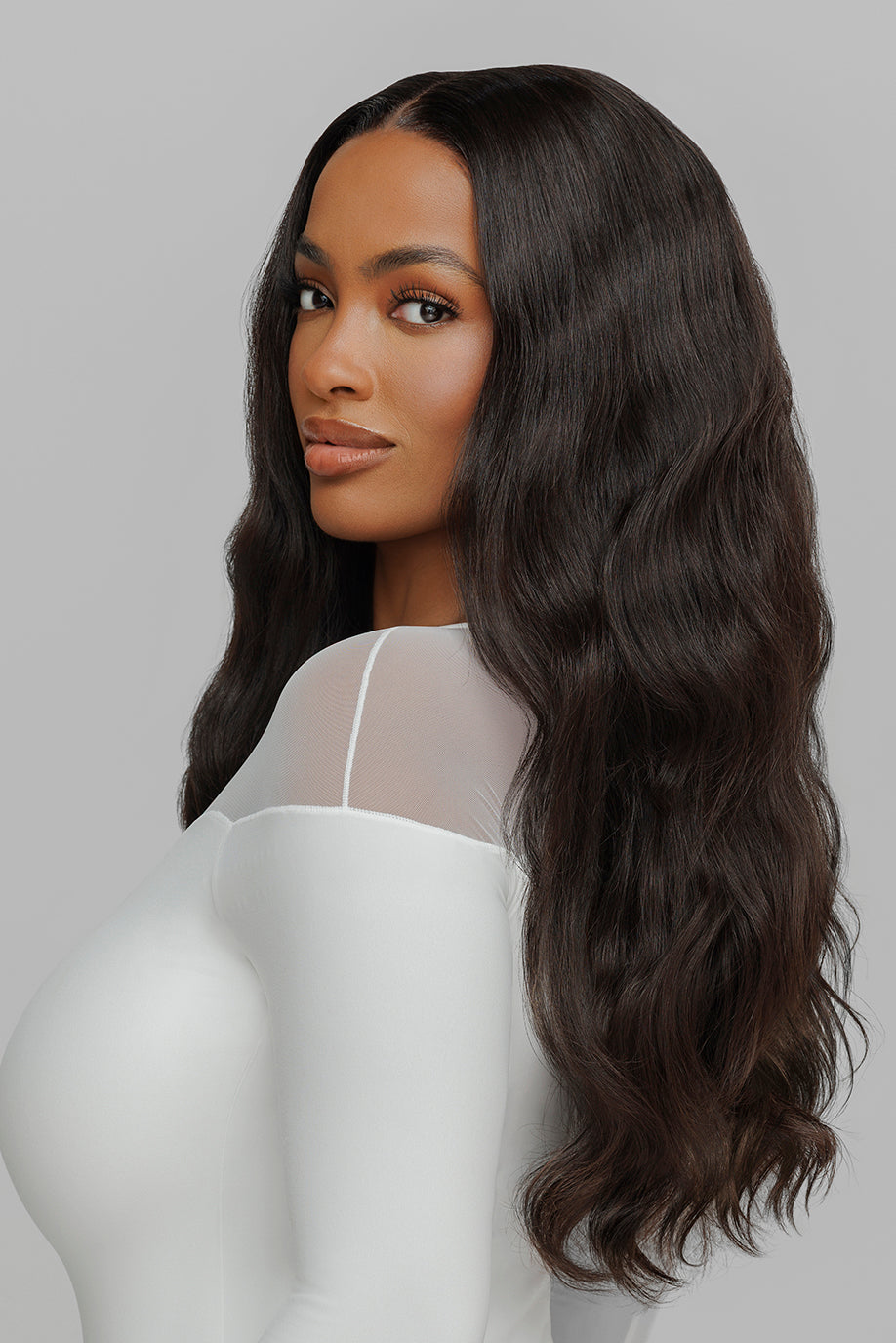 Woman with long, wavy hair wearing a white top, looking over her shoulder.