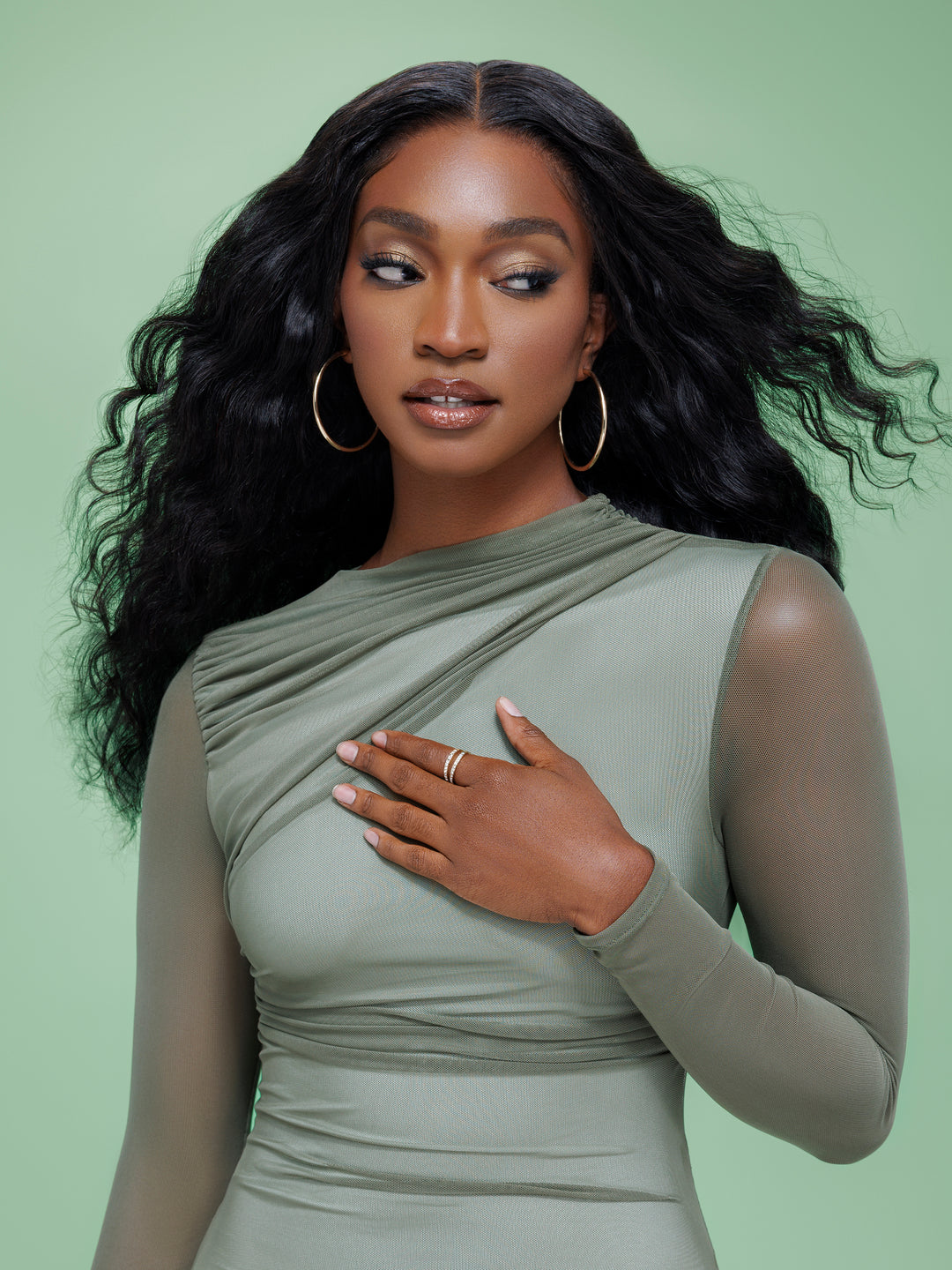 Black woman with long wavy hair looking away, wearing a sage green mesh top and gold hoop earrings.