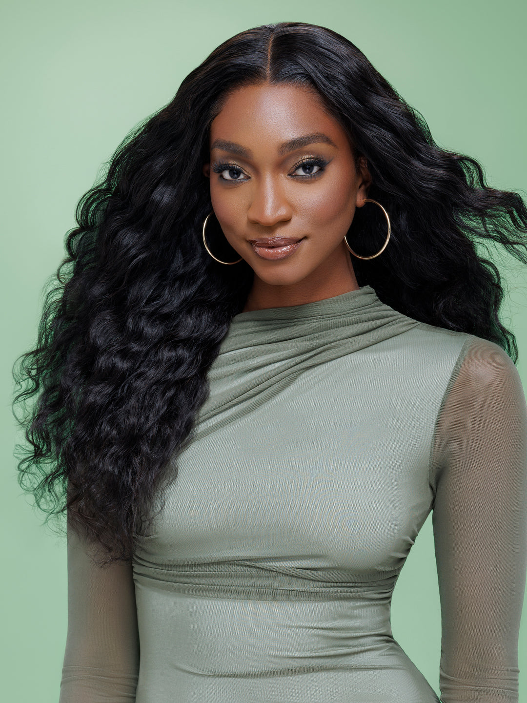 Black woman with long wavy dark hair wearing a sage green mesh top and gold hoop earrings.